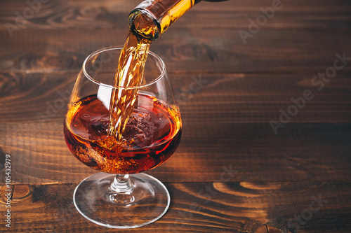 Pouring brandy or cognac from the bottle into the glass against wooden background photo