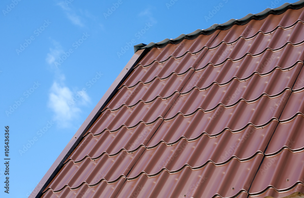 Part of country house roof from brown metal tile under blue sky with white clouds. Horizontal view closeup