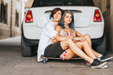 Couple hugging each other sitting on the skateboard desk in front of the car 