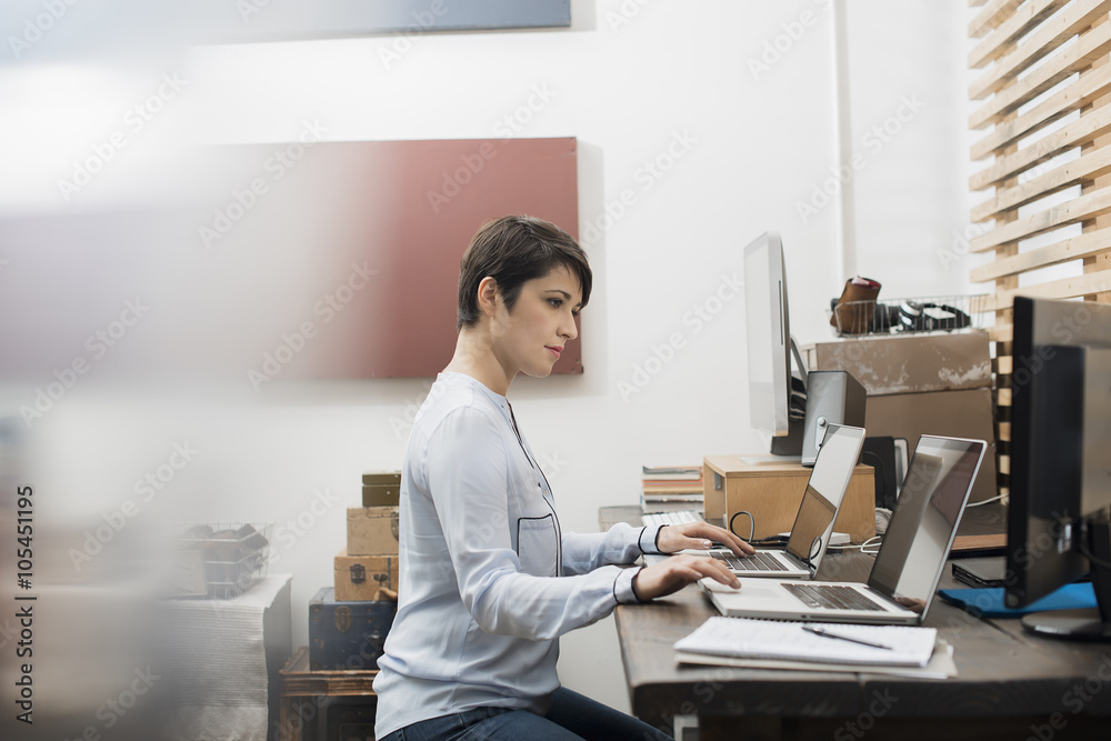 A woman in a home office seated at a desk with two laptops, her hands on the keyboard of one computer, looking at the screen on the other, A woman in a home office seated at a desk with two laptops, her hands on the keyboard of one computer, looking at the screen on the other,