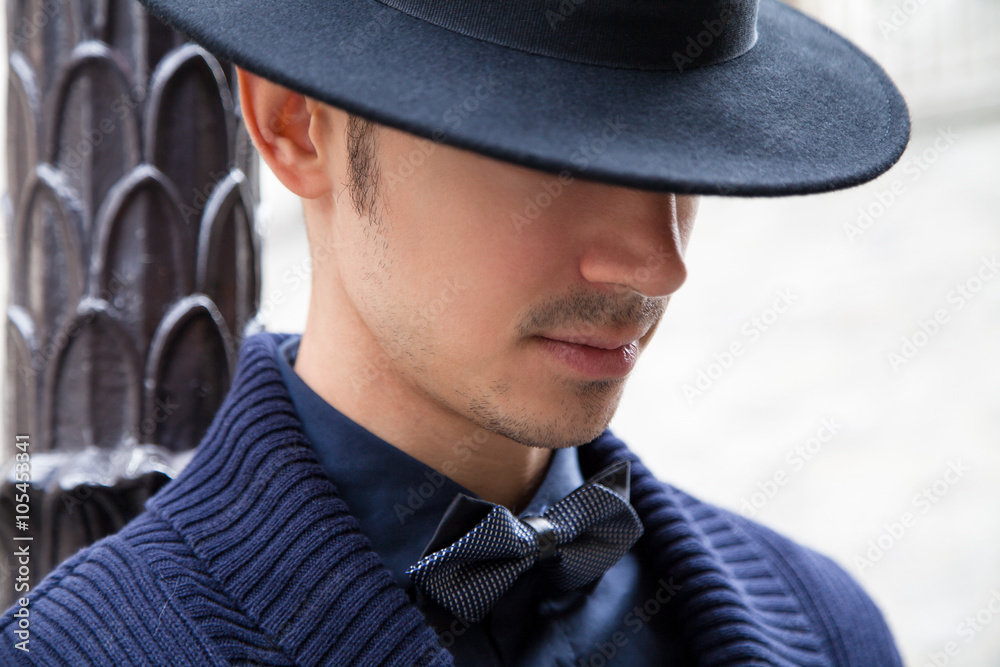 Young handsome man wearing a bow tie and a hat on her head. Elegant male model posing outdoor. Close up picture of an attractive guy
