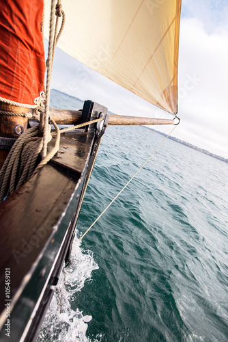 bow of a ship with a beige cotton jib sail filled by the wind with wooden mast, bowsprit and hull of an old rigging sailing boat during a sunny sea trip in brittany with a blur background