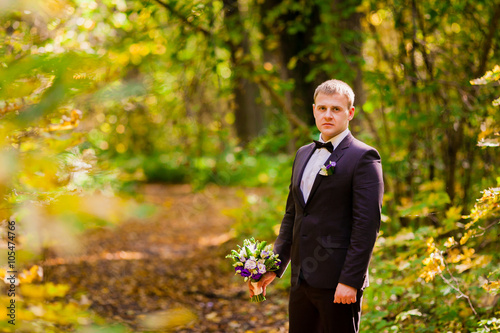Жених стоит в осеннем лесу на фоне желтых листьев и держит букет невесты