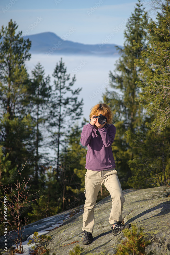 Young female photographer standing on the top of the mountain on big rock and taking picture. Front view. Trees and mountain on the background.