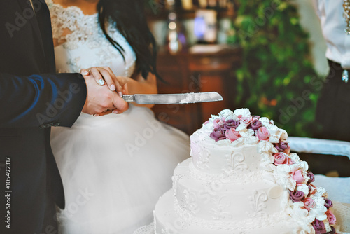 A bride and a groom is cutting their wedding cake with purple and white flowers