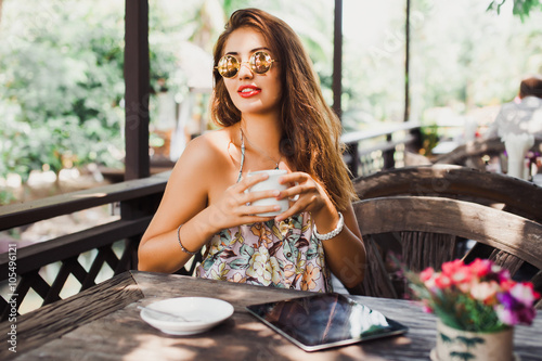 beautiful young woman in sunglasses sitting in a cafe drinking coffee/tea and work on your tablet on the Internet, outdoor portrait, close up 