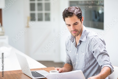 Young man reading documents while sitting at desk with laptop 