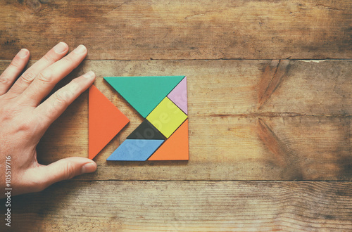 man's hand holding a missing piece in a square tangram puzzle, over wooden table.
 photo