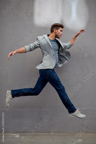 Stylish man in blue jacket jumping opposite grey wall