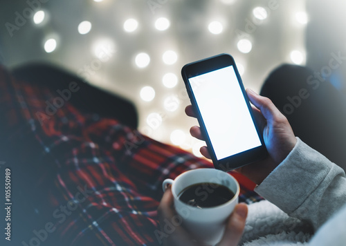 Girl pointing finger a black smartphone with a empty blank screen monitor and a cup of coffee or tea on the background bokeh light in a homely atmosphere, smart phone with space for information