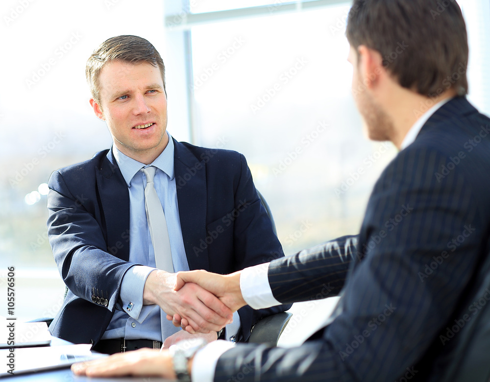 Business colleagues sitting at a table during a meeting with two male executives shaking hands Business colleagues sitting at a table during a meeting with two male executives shaking hands