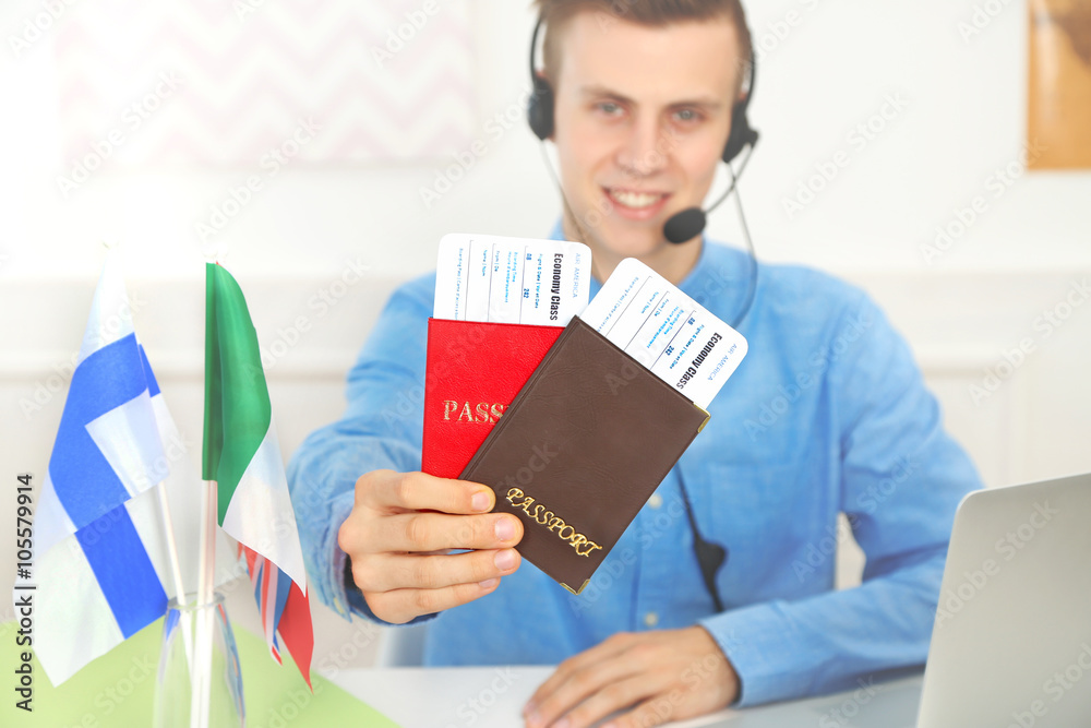 Young man talking on headset and showing passports with tickets in the office of travel company Young man talking on headset and showing passports with tickets in the office of travel company