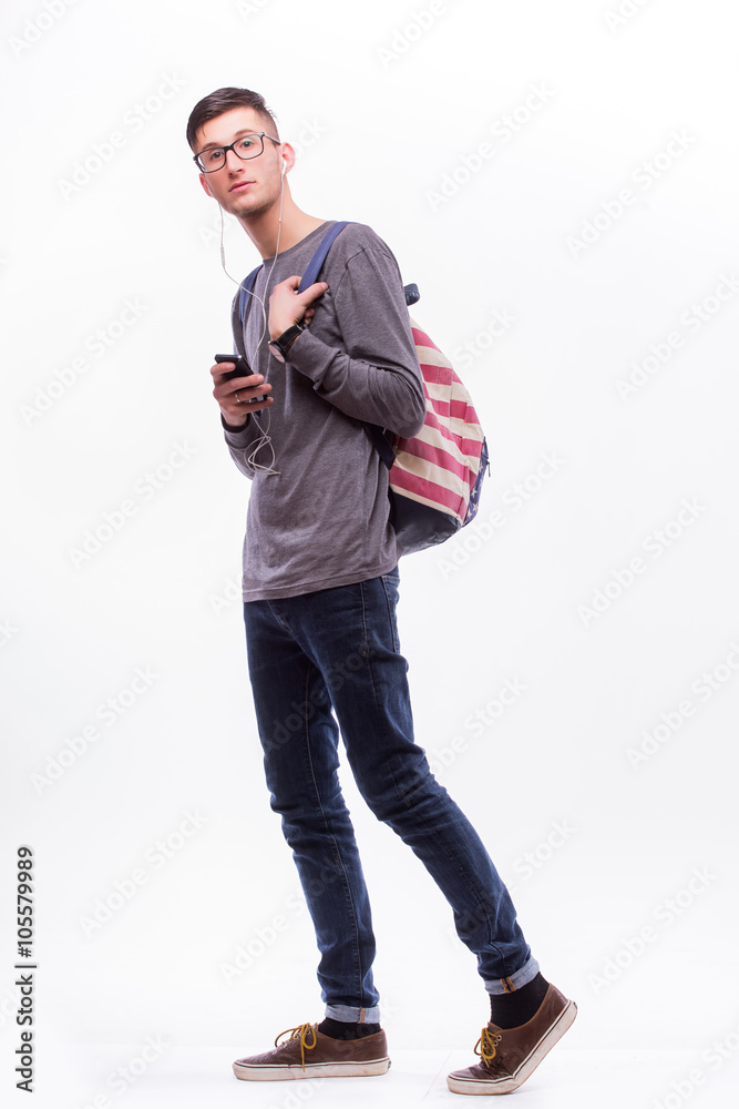 Fototapeta premium Happy hipster guy in glasses with backpack using a smart phone to listen music with headphones on white background