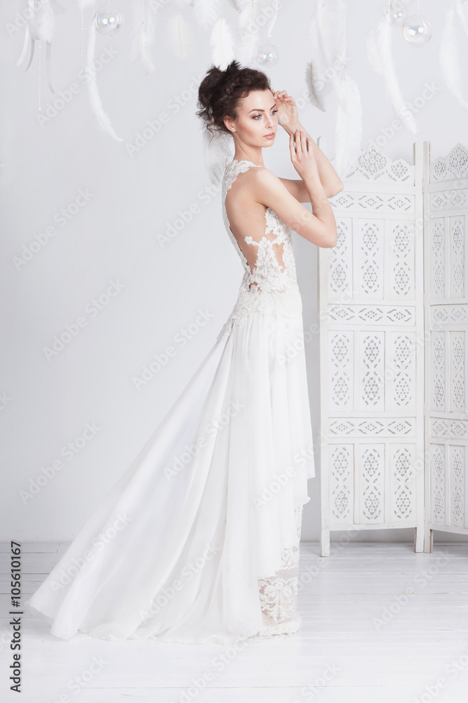 Tall and slim young bride in a luxurious lace wedding dress. She stands  with her back to the viewer. She has dark hair. White feathers around her.  Stock Photo | Adobe Stock