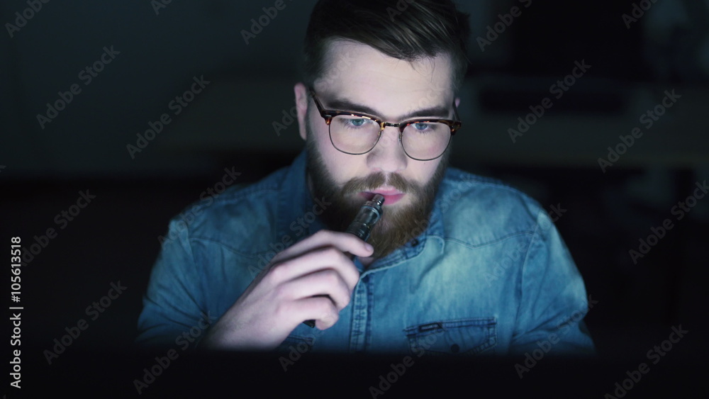 Casual man smoking electronic cigarette and using PC at night Casual man smoking electronic cigarette and using PC at night