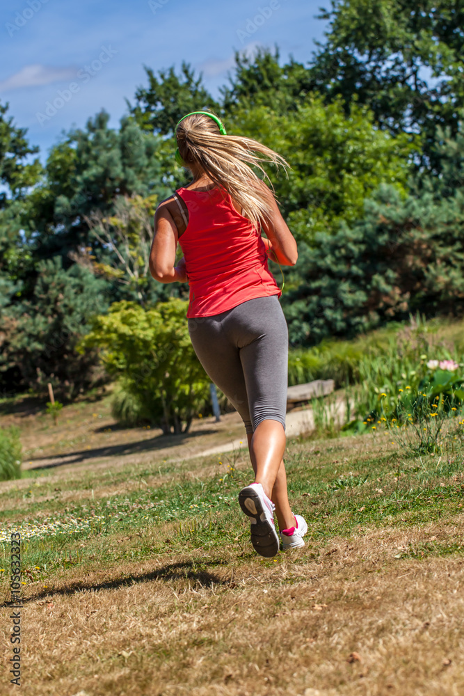 outdoors sports - back view of 20s blond woman jogging with green headphones on, listening to music in park, sunny day outdoors sports - back view of 20s blond woman jogging with green headphones on, listening to music in park, sunny day