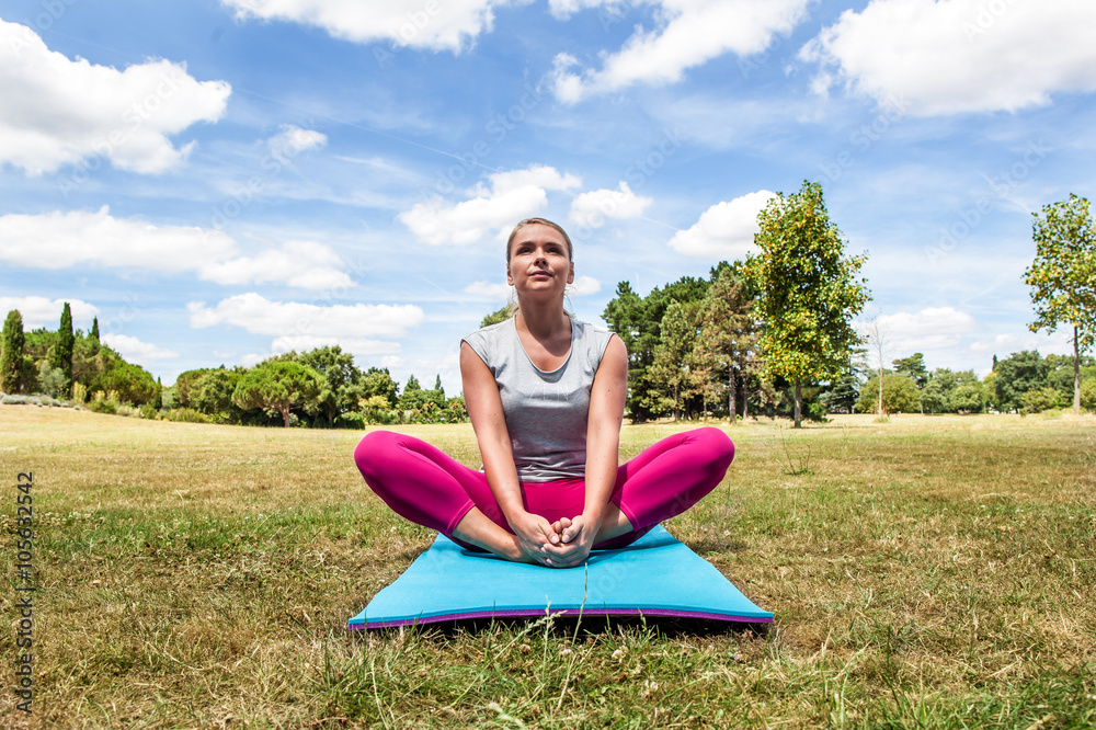 wellbeing and stretching outside - relaxed young blond woman doing yoga with tight legs and upper body on exercise mat over summer blue sky