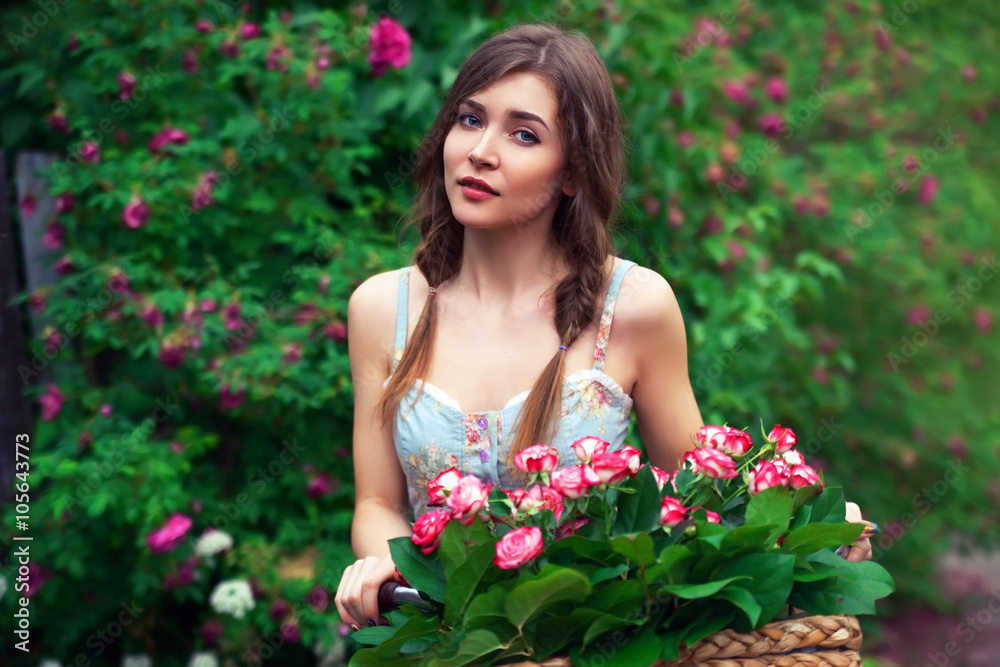 Beautiful girl with bicycle and basket full of roses on floral background. Vintage style.