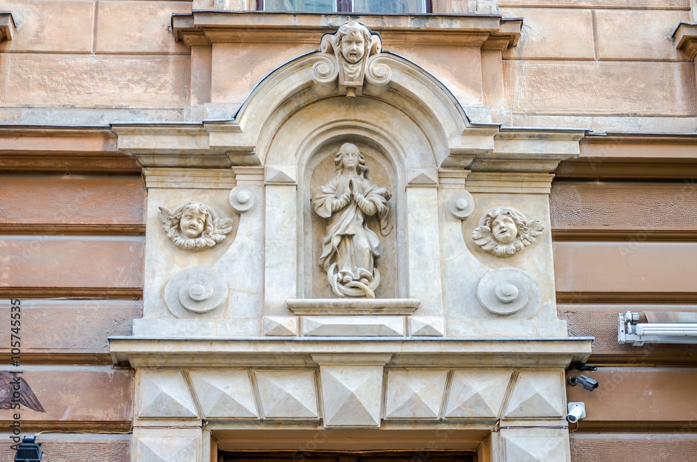 Religious statue of holy woman who prays with her hands folded, surrounded by heads of angels who look at it, on the facade of one of the historic buildings on the street of Lviv, Ukraine