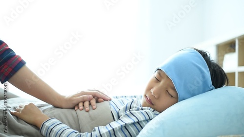 Sick boy sleeping on the bed with cooler gel on his head. His mother check thermometer for the body temperature.