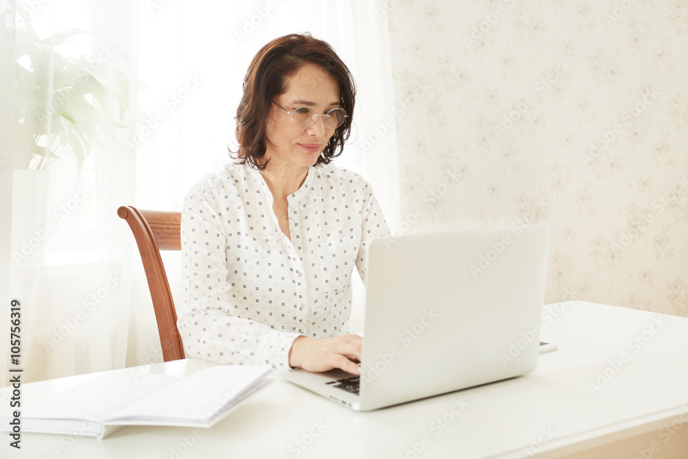 Beautiful asian mature female using portable laptop computer while work at compilation of business charts, woman sitting at the table with open net-book in office or home interior. Beautiful asian mature female using portable laptop computer while work at compilation of business charts, woman sitting at the table with open net-book in office or home interior.