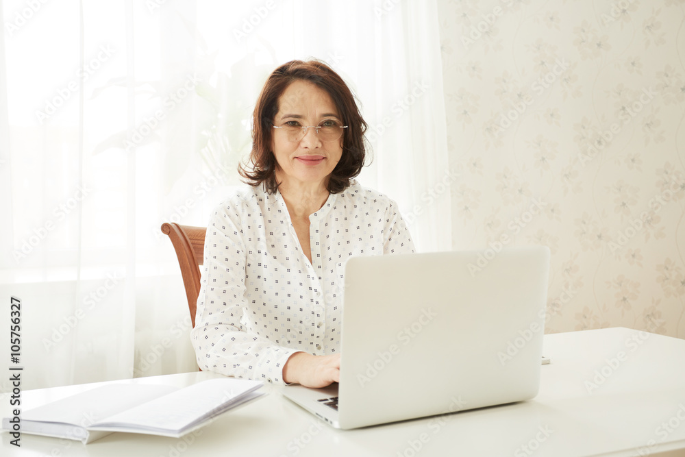 Beautiful asian mature female using portable laptop computer while work at compilation of business charts, woman sitting at the table with open net-book in office or home interior. Beautiful asian mature female using portable laptop computer while work at compilation of business charts, woman sitting at the table with open net-book in office or home interior.