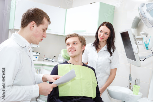 Cheerful young dentist is examining teeth of young man. He is holding patient card.  The female assistant is halping