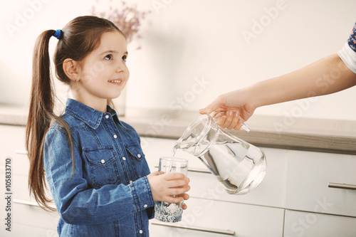 фотографія Little girl holding glass and adult woman pouring water from jug in living room