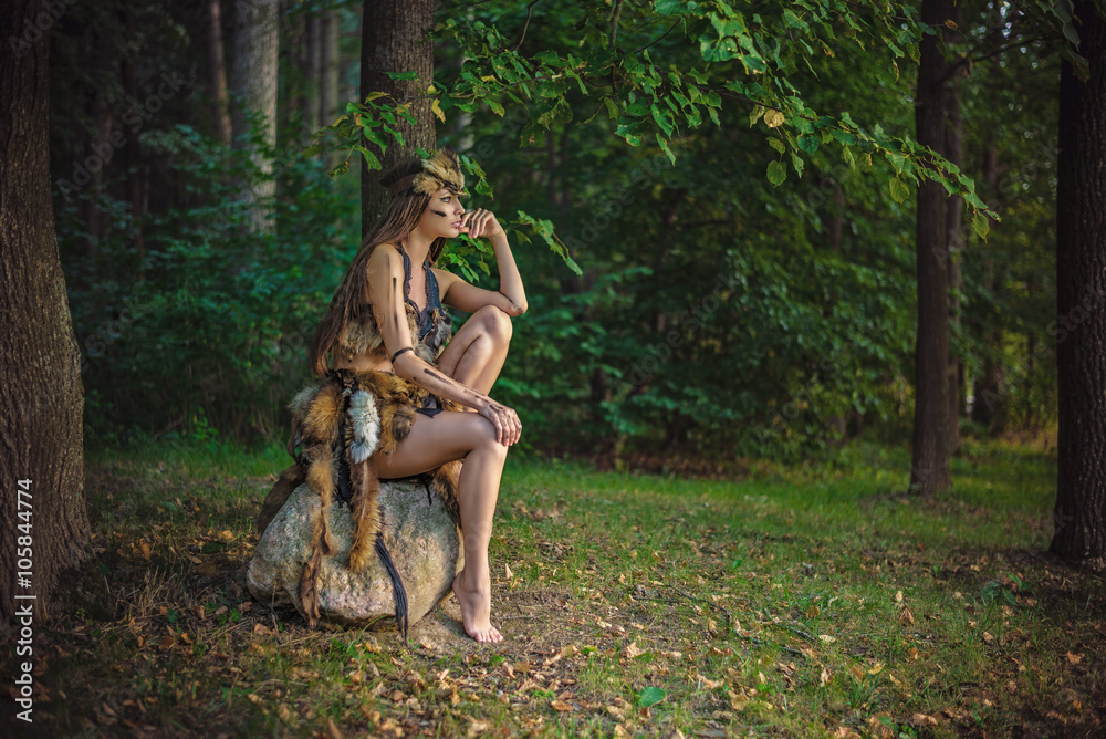 Fototapeta premium Elegancka piękna młoda Amazonka długowłosa i długonoga siedząca na skale w lesie