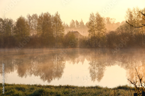 Foggy landscape: early morning on the river in the spring, the silhouette of wooden house over the water. Village, countryside. Fog. Sun.