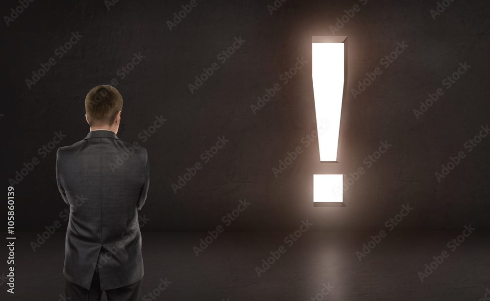 Obraz premium Businessman standing in front of concrete wall with bright big exclamation sign. Financial and business concept.