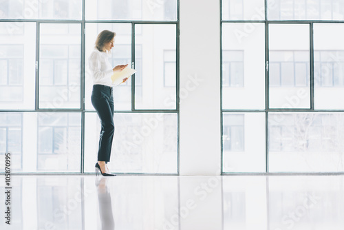 Photo business woman wearing modern suit, looking mobile phone and holding papers in hands. Open space loft office. Panoramic windows background. Horizontal mockup.
