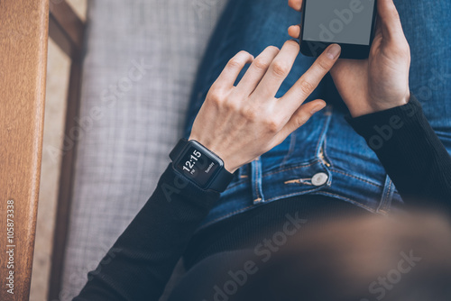 Photo of female hands touching screen smartphone and wearing generic design smart watch. Film effects, blurred background. Horizontal