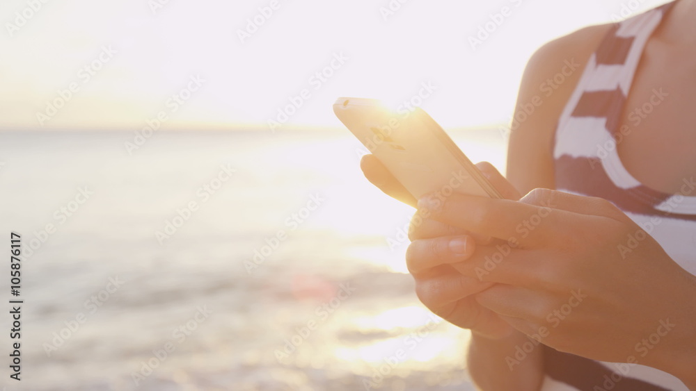 Woman texting on smartphone. Female is using mobile cell smart phone app at beach during sunset. Waves are rushing towards her. Sun is shining in background.