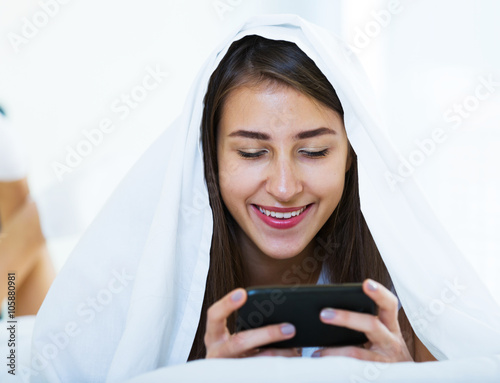 Smiling girl with smartphone hiding in bed photo