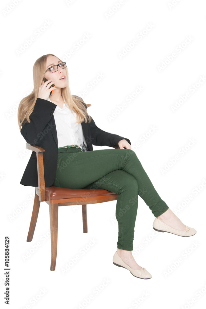Attractive young woman in glasses dressed in white blouse, black jacket and green jeans sitting in a chair, talking on cell phone and looking up (isolated on white background) Attractive young woman in glasses dressed in white blouse, black jacket and green jeans sitting in a chair, talking on cell phone and looking up (isolated on white background)