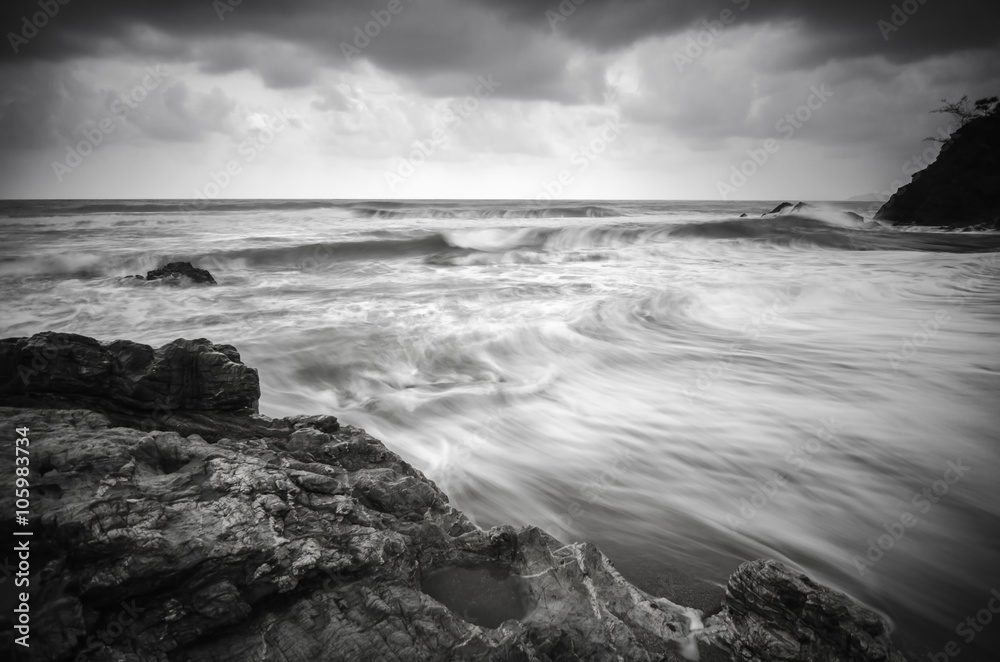 black and white image wave flow on the sandy and rocky beach with dark and dramatic cloud.
