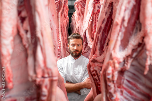 Fotografija Portrait of a handsome butcher in white uniform with pork carcasses at the meat