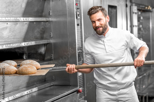 Handsome baker in uniform taking out with shovel freshly baked buckweat bread from the oven at the manufacturing photo