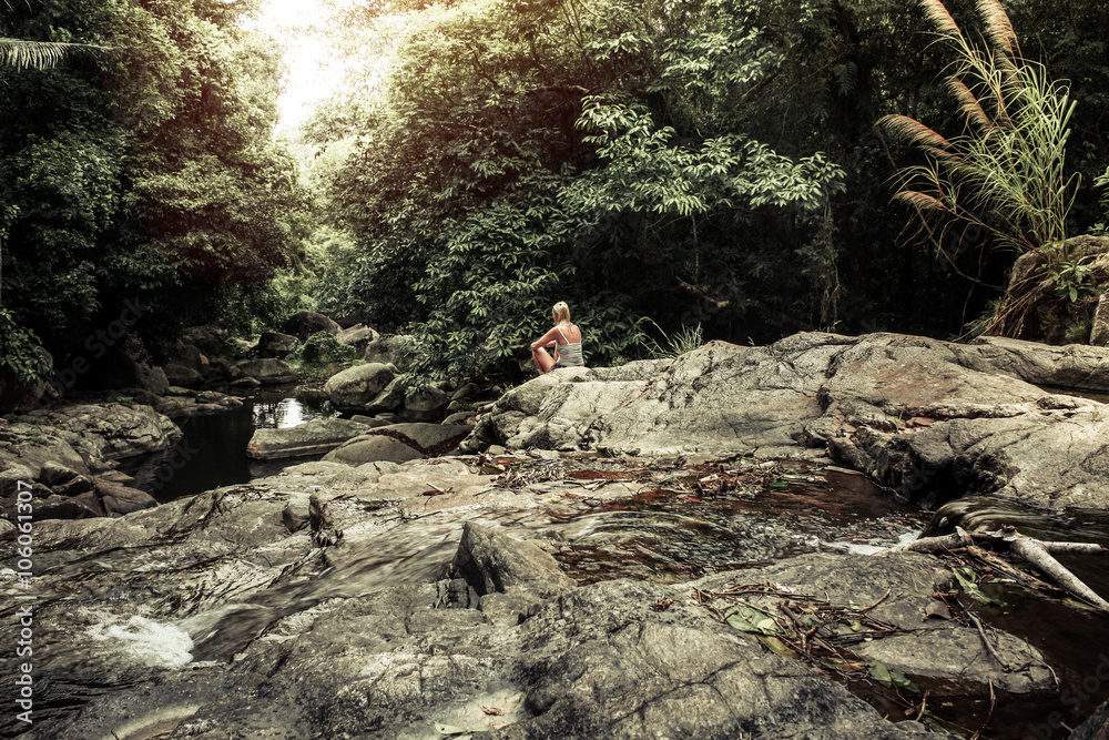 meditating young woman enjoying the pristine beauty of nature in tropical rainforest at waterfall  during sunset far away from bustle. Rays of light breaking through the dense foliage