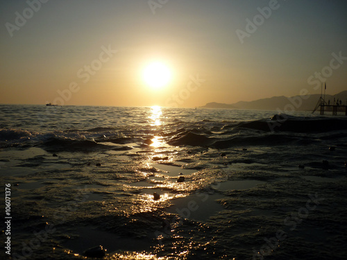 Marine sunset scene with lights reflecting at dark seaways in foreground and sun with halo glade shines over horizon in dusk against mountains silhouette with clouds background