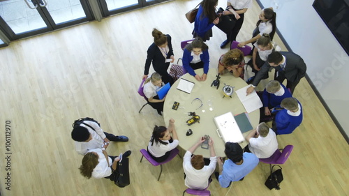 View from above of students and teachers in a technology lesson. Some are sitting at the table whilst others are standing and talking amongst themselves.