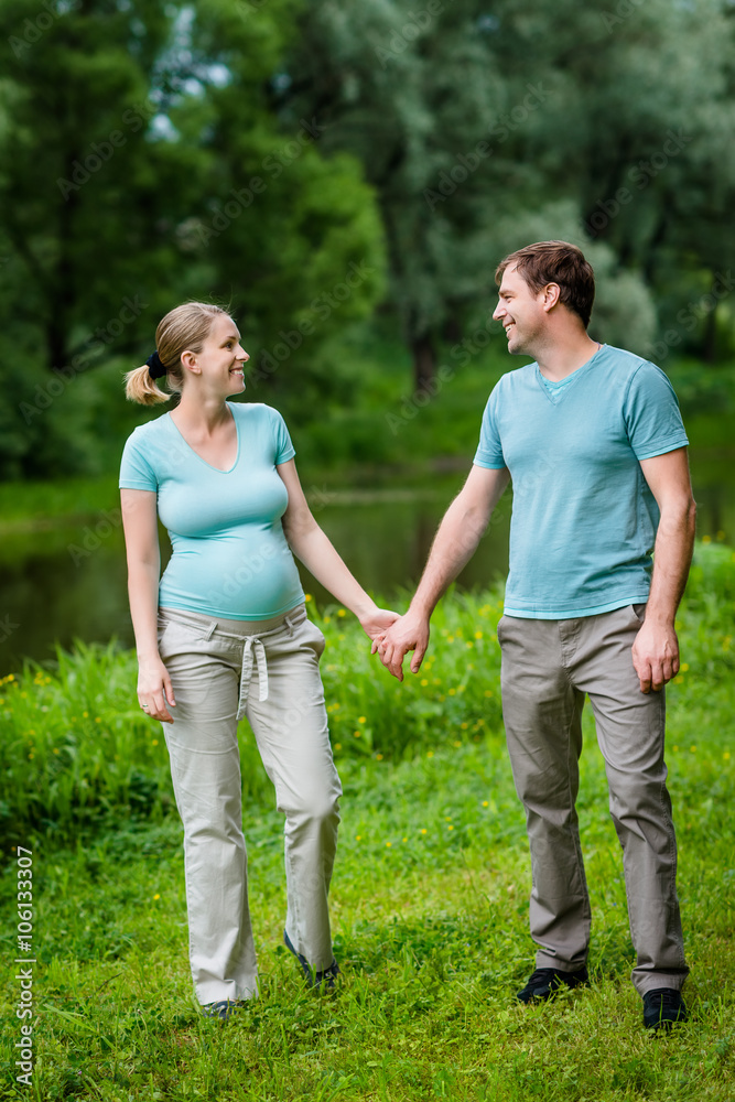 Adorable young pregnant woman and happy handsome man smiling, holding each other hands and looking at each other in summer park. Happy family and pregnancy concept. Mother's Day