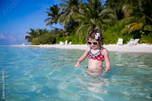 Cute little girl in bathing suit playing in the clear ocean on the tropical resort beach with white sand, azure blue sea and coconut palms. Paradise Landscape background. Happy and excited child. 