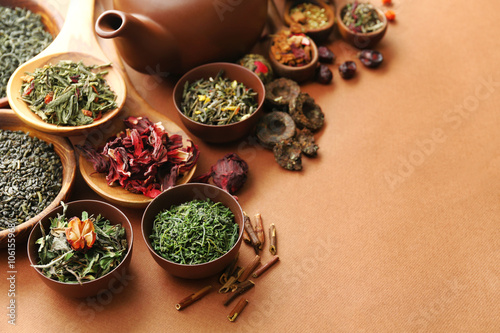 Variety of dry tea in bowls with teapot on light brown background, closeup