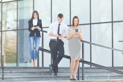 Three busy business people at break at work. Young businessman and two businesswomen are using their mobile phones while walking down the stairs in front of the modern office building.