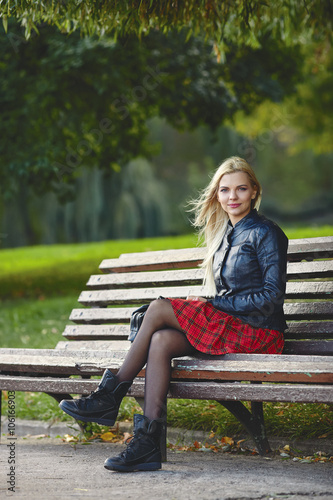 Closeup outdoors portrait of young adorable blonde woman sitting on the park bench in windy weather conditions with green blurry background