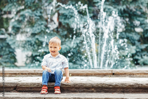 Portrait of cute adorable funny Caucasian little boy toddler in white shirt and blue jeans playing laughing smiling having fun by fountain in summer outside