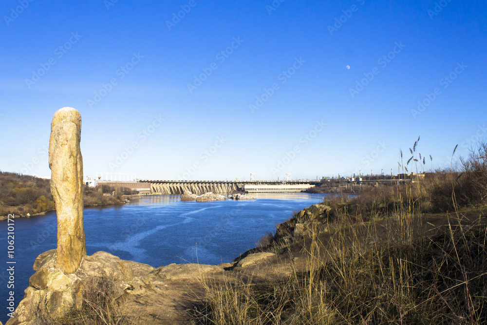 conceptual picture - a combination of ancient and modern. Stone idol statue on a rock above the river. Horizontal shot, landscape.