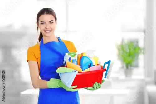 Woman in rubber gloves holding basin with detergents on blurred background